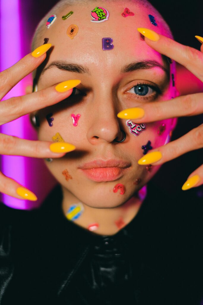 Crop unemotional bald female with multicolored stickers on face skin touching face and looking at camera in dark studio